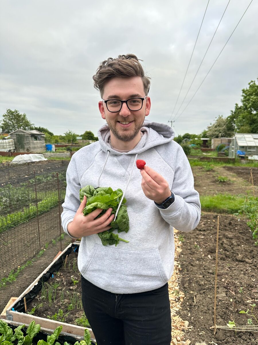 Calum at the allotment holding freshly picked spinach and a strawberry